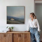 A woman in a white blouse and jeans leans on a wooden cabinet, gazing at the Rustic Solitude Canvas Art - Square, which depicts a distant house and rolling hills. A plant and books sit neatly atop the cabinet.
