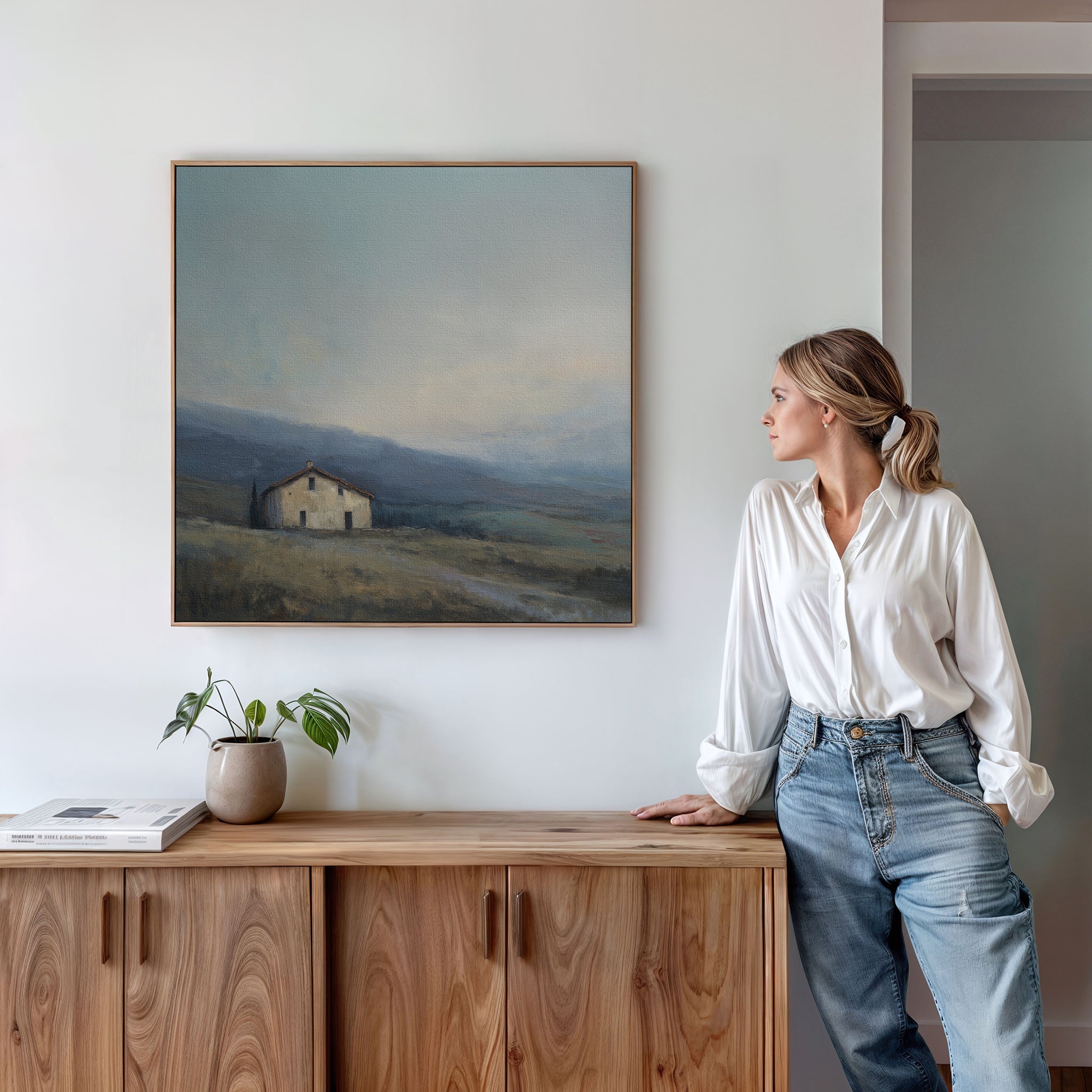 A woman in a white blouse and jeans leans on a wooden cabinet, gazing at the Rustic Solitude Canvas Art - Square, which depicts a distant house and rolling hills. A plant and books sit neatly atop the cabinet.