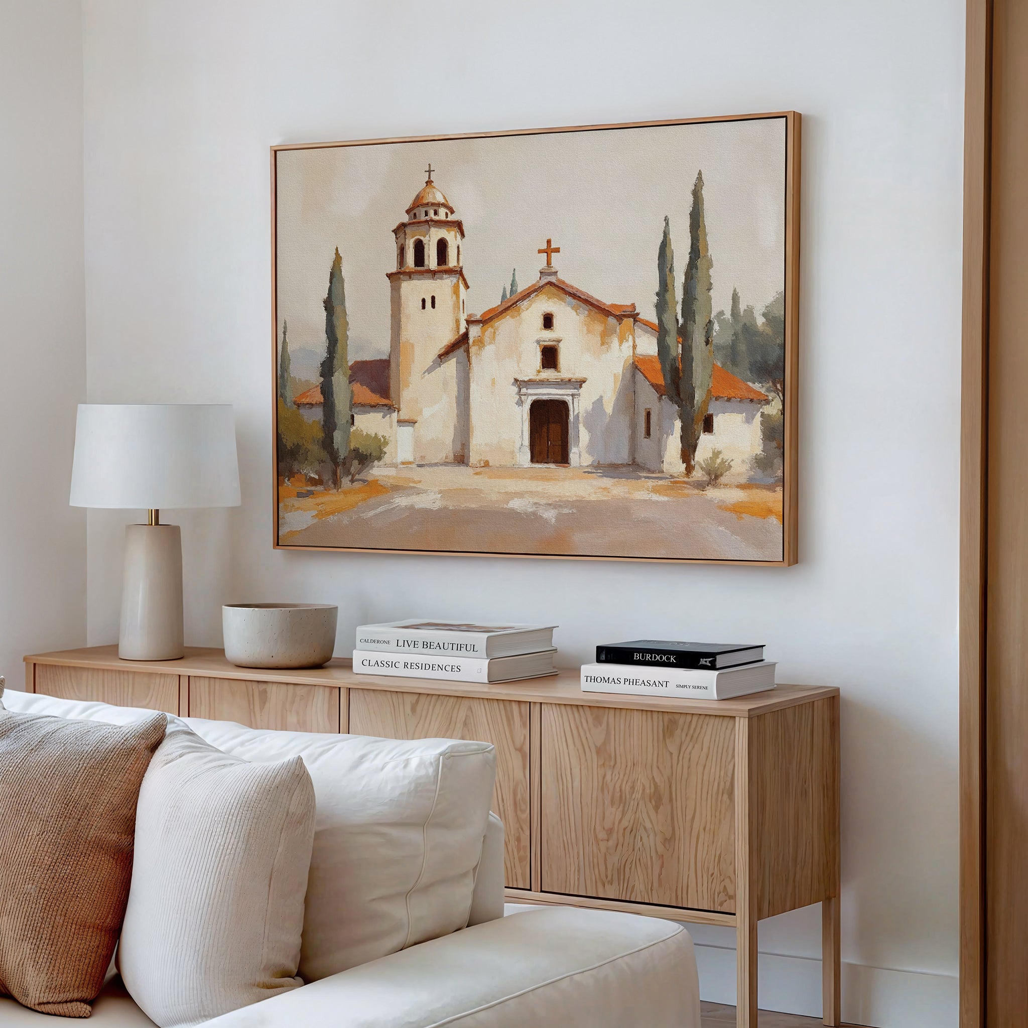 A cozy living room features a wooden console table with books, a lamp, and a ceramic bowl. Above the white sofa and pillows hangs the Sacred Courtyard Canvas Art - Horizontal, adding charm to the space.