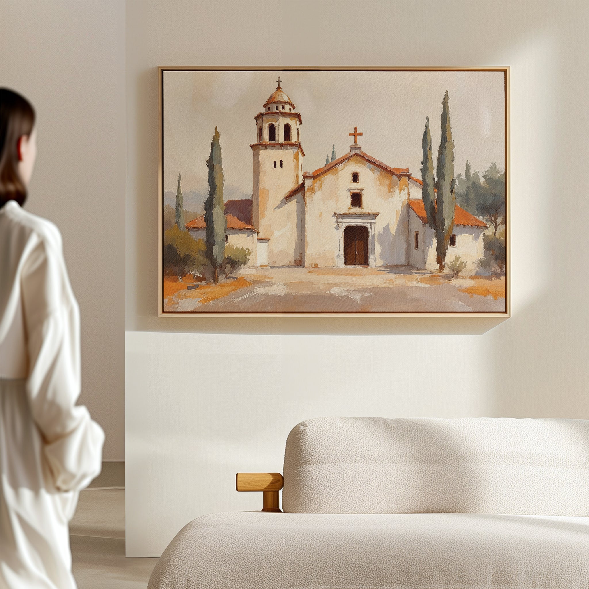 A woman stands in a softly lit room, gazing at the Sacred Courtyard Canvas Art - Horizontal, which is hung on a light-colored wall above a beige sofa and depicts a Spanish colonial church with two bell towers and cypress trees.