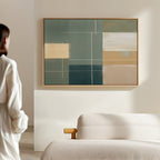 A woman in a white blouse stands by a light sofa, admiring the Sage Compartments Canvas Art - Horizontal—an abstract piece with geometric shapes in muted green, beige, cream, and blue tones that elevates the modern living room decor.