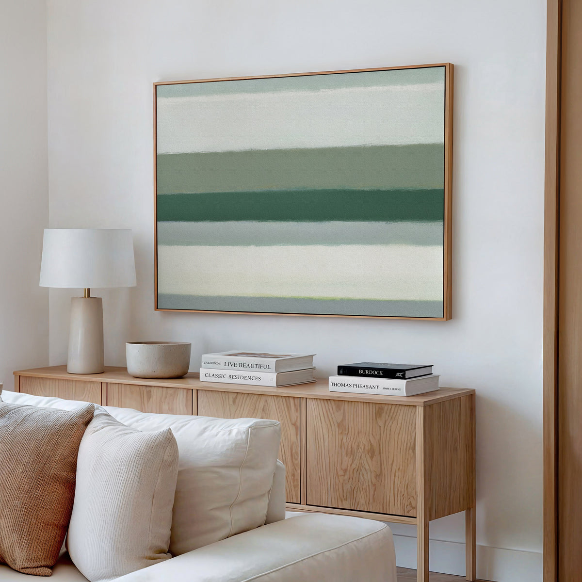 A modern living room with a light wood sideboard, white lamp, decorative bowl, stacked books, and the Sage Gradient Canvas Art - Horizontal featuring abstract green and beige stripes on a white wall.