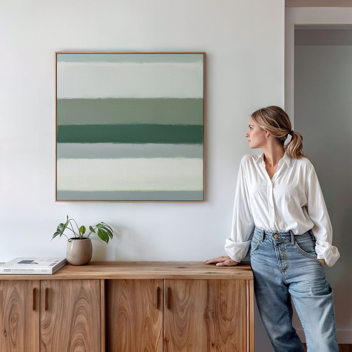 A woman in a white blouse and jeans leans against a wooden cabinet, looking at the Sage Gradient Canvas Art - Square on a white wall. A potted plant and a book sit atop the cabinet.