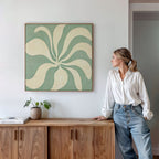 A woman in a white blouse and jeans leans against a wooden cabinet, gazing at the Sage Meditation Canvas Art - Square in sage green above her. A potted plant and books on the cabinet complement the Nordic Park-inspired decor.