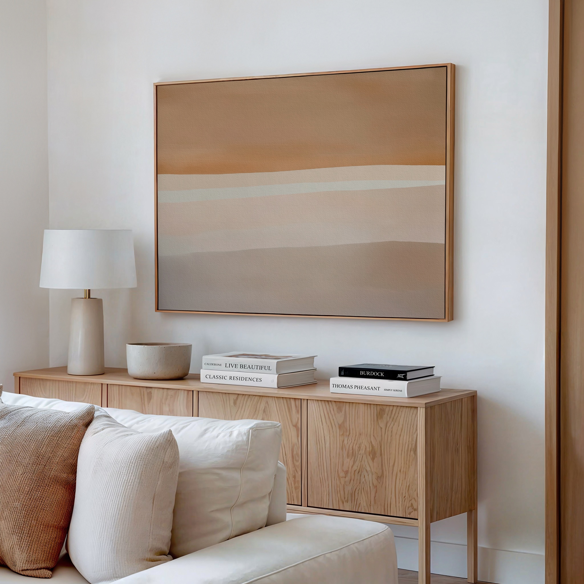 A minimalist living room features a cream-colored sofa, light wood sideboard, stacked books, white lamp, decorative bowl, and Saharan Lines Abstract Canvas Art in beige and earth tones on the white wall above.
