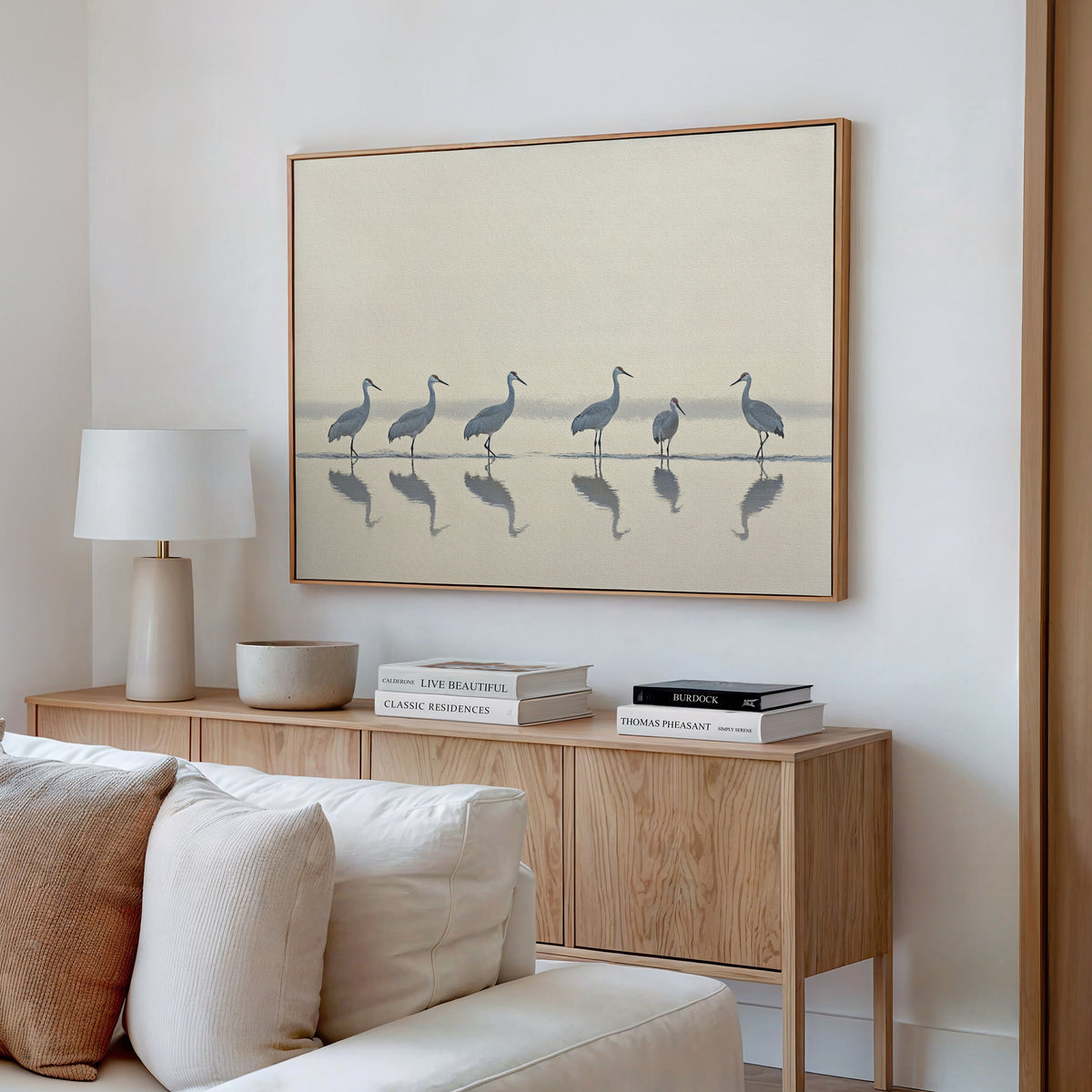 A minimal living room features a light wood console, books, a ceramic lamp, and the Sandhill Procession Canvas Art - Horizontal, showing elegant cranes in shallow water. Neutral colors and soft natural light enhance the tranquil ambiance.