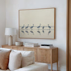 A minimal living room features a light wood console, books, a ceramic lamp, and the Sandhill Procession Canvas Art - Horizontal, showing elegant cranes in shallow water. Neutral colors and soft natural light enhance the tranquil ambiance.