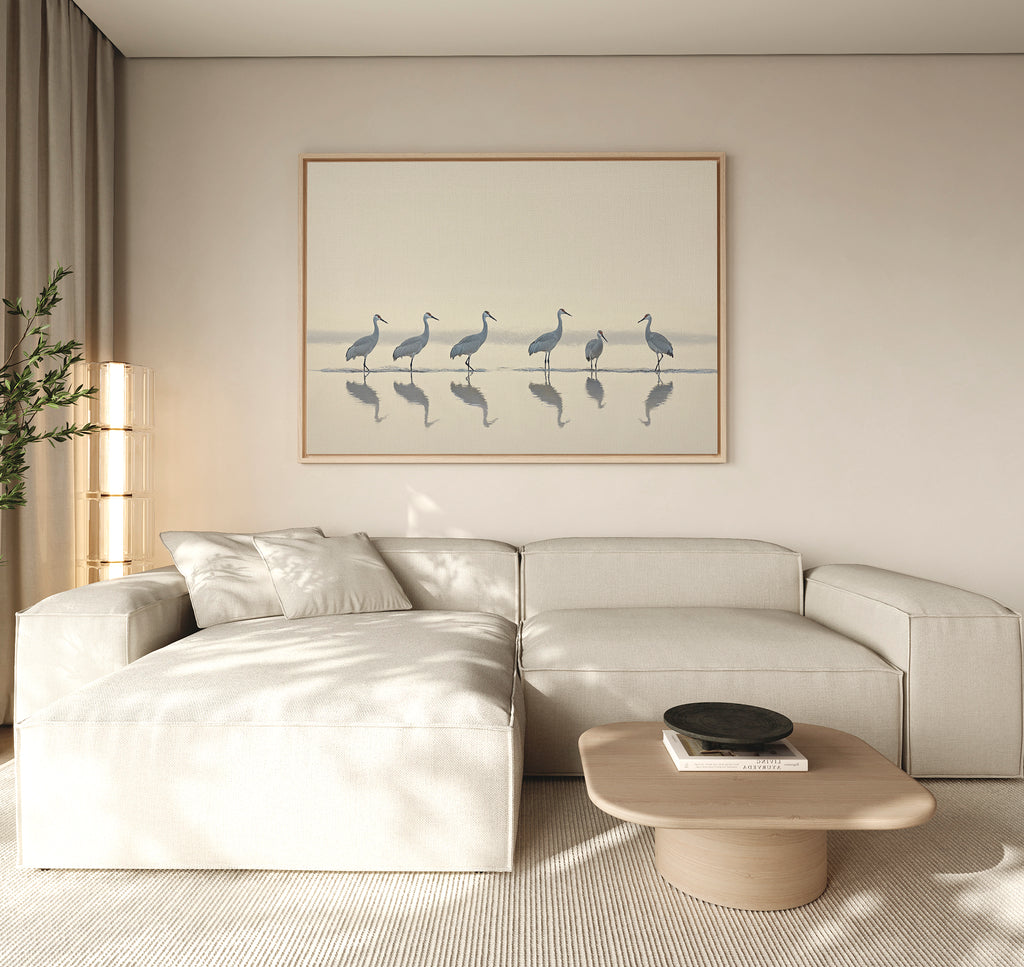 A modern living room with neutral tones, a beige sectional sofa, wooden coffee table, potted plant, and the Sandhill Procession Canvas Art - Horizontal featuring elegant cranes in water. Soft natural light creates a tranquil ambiance.