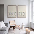 A modern living room with neutral tones features a wooden chair, a small white table with a lit candle, serene home decor, and large windows. Above the light sofa hangs Sandhill Procession - Set Of 2 artwork.
