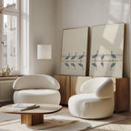 A modern living room features two rounded white chairs, a low wooden table, and a wooden sideboard topped with Sandhill Procession - Set Of 2 artwork. Sunlight pours through large windows, creating a bright and serene home decor ambience.