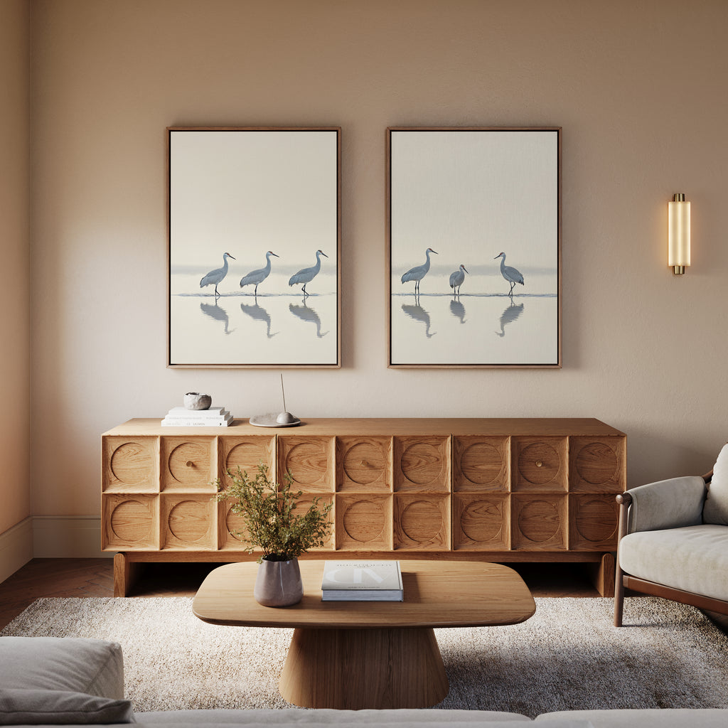 A cozy living room with a wooden sideboard, coffee table, beige armchairs, and serene decor. The Sandhill Procession - Set Of 2 framed prints hang on the wall, while a small vase with flowers and a modern wall light complete the look.