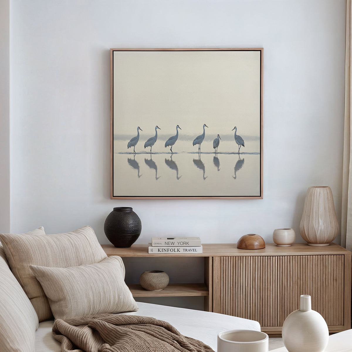A modern, minimalist living room features neutral decor with a light sofa, vases, and books on a wooden sideboard. Above hangs the Sandhill Procession Canvas Art - Square, adding tranquility and elegance to the space.