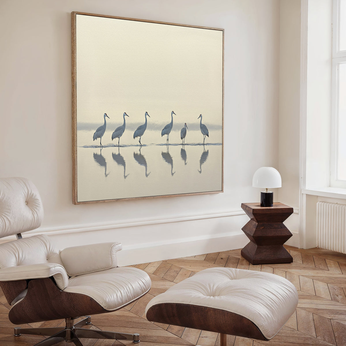 A modern living room features a white lounge chair, matching ottoman, wooden side table with a white lamp, and the Sandhill Procession Canvas Art - Square depicting six birds by water. Light from a tall window adds to the serene, inviting atmosphere.