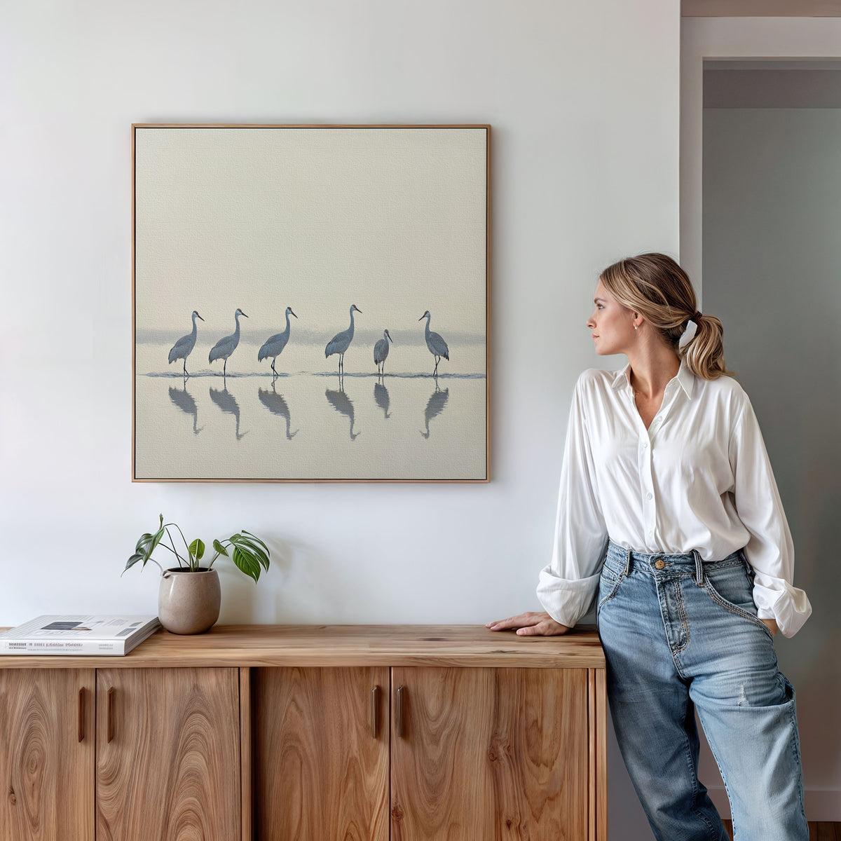 A woman in a white blouse and jeans stands by a wooden cabinet, admiring the Sandhill Procession Canvas Art - Square, featuring tranquil cranes reflected in water. A small plant and books on the cabinet complete the peaceful Nordic-inspired look.