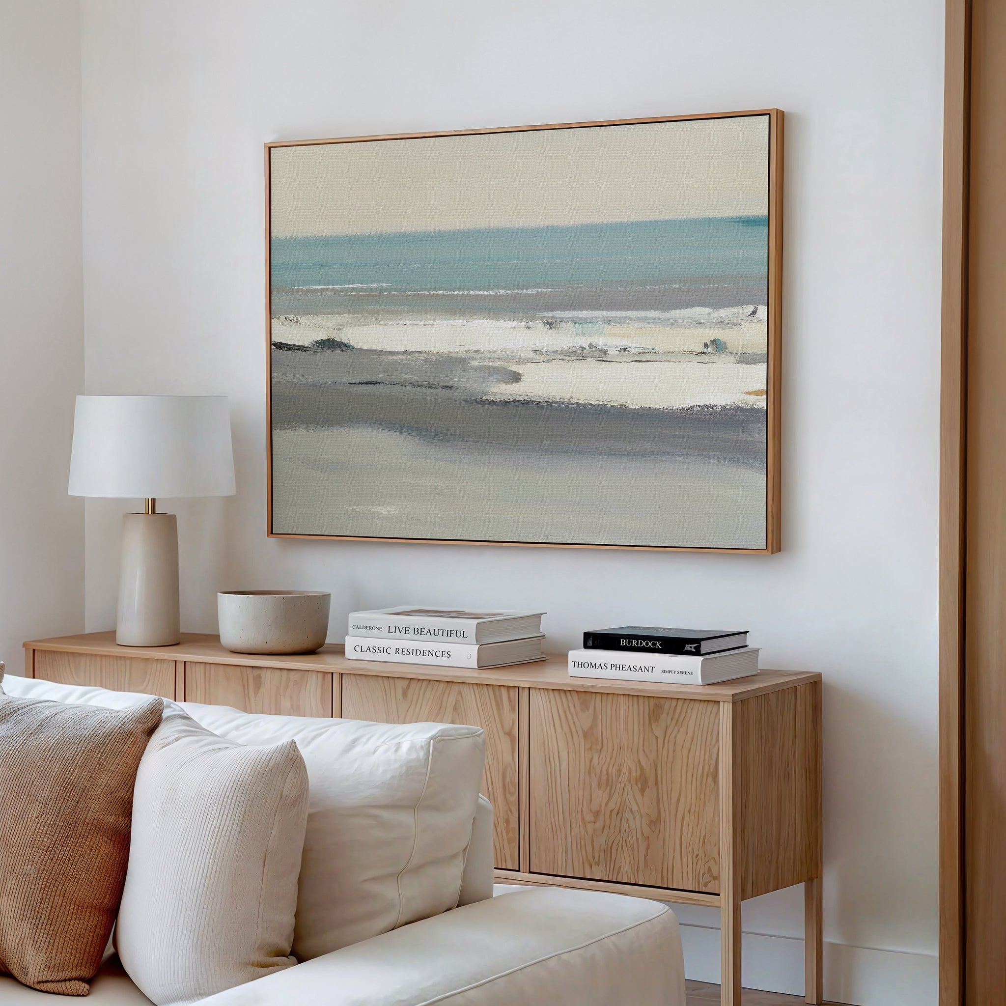 A modern living room features a wooden sideboard with stacked books, a white table lamp, a ceramic bowl, and the Sandy Retreat Canvas Art - Horizontal hanging above a beige sofa on a white wall.