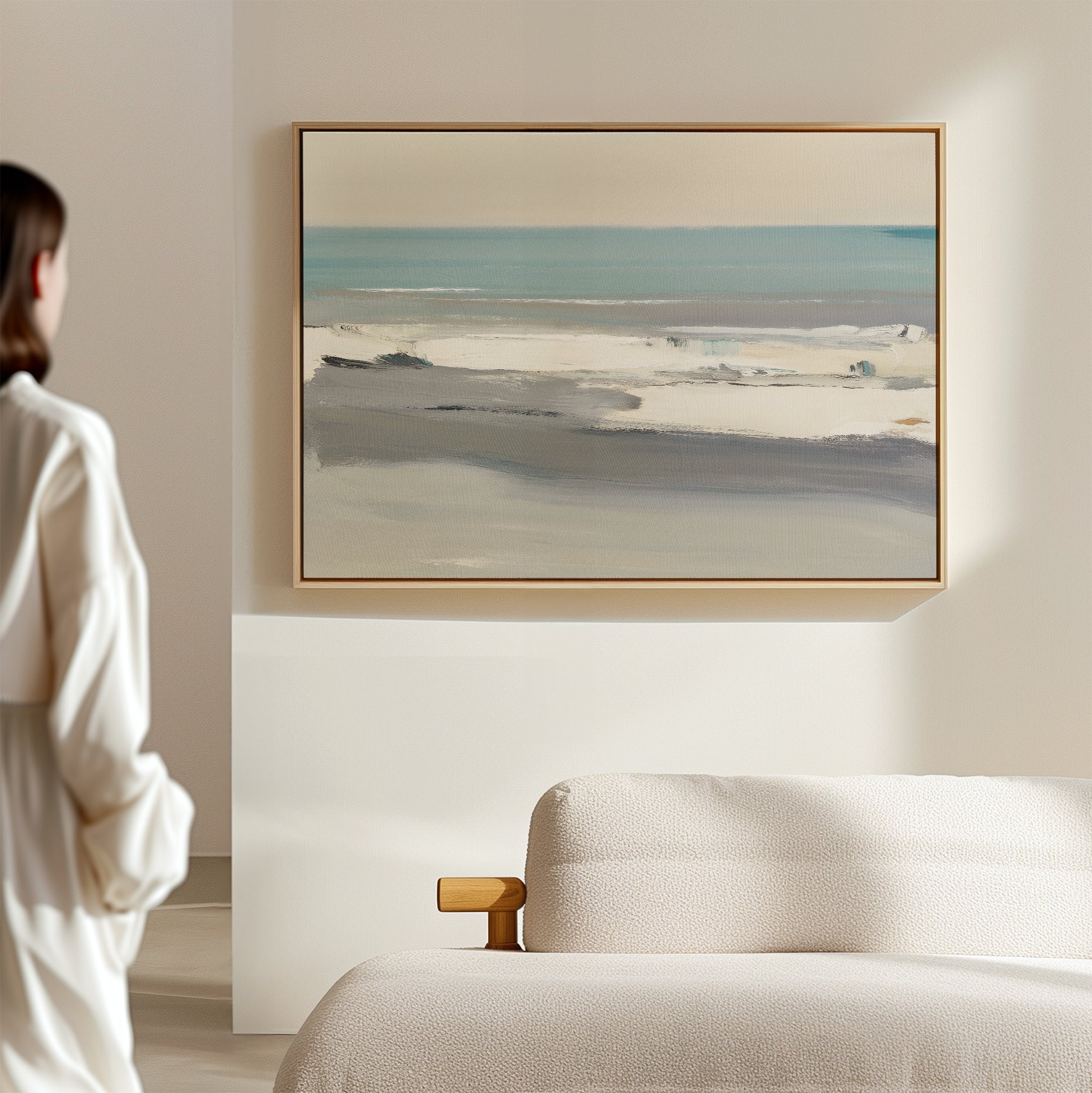 A person in a white outfit stands near a bed, admiring the Sandy Retreat Canvas Art - Horizontal on the wall, its soft blue, beige, and gray tones adding subtle seaside vibes to the minimal, modern room.