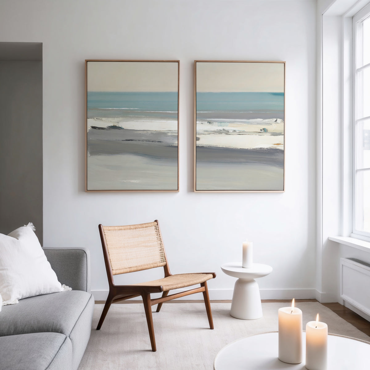 A modern, minimalist living room features a woven chair, round side table, and candles. Two large "Sandy Retreat - Set Of 2" coastal ocean prints hang on a white wall above, with natural light streaming through a big window.