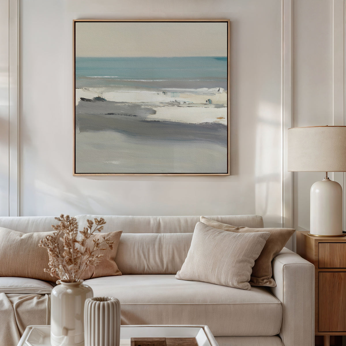 A neutral-toned living room features a beige sofa with matching pillows and a dried floral arrangement. Above, the Sandy Retreat Canvas Art - Square adds gentle seaside vibes with its serene blue and gray coastal scene.