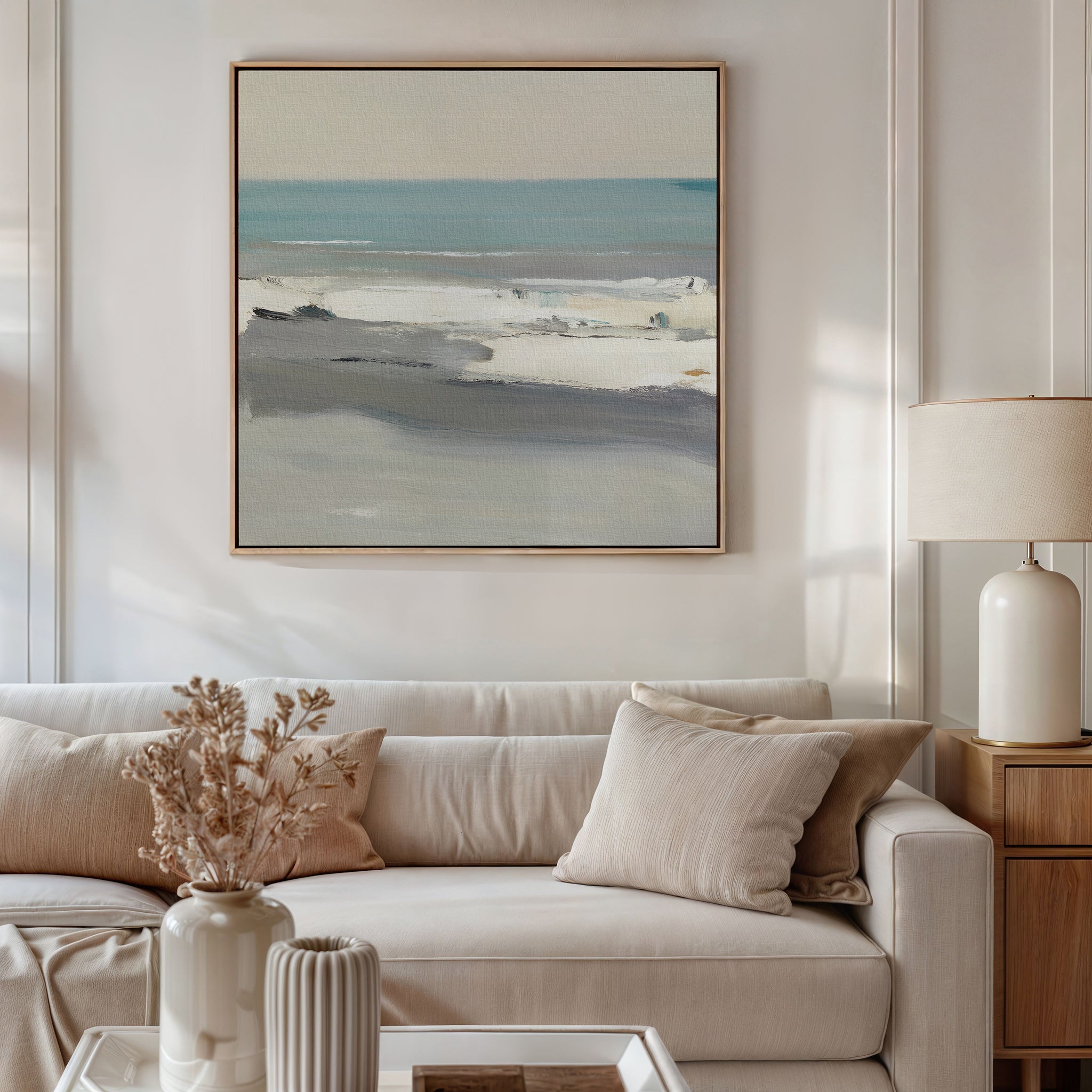 A neutral-toned living room features a beige sofa with matching pillows and a dried floral arrangement. Above, the Sandy Retreat Canvas Art - Square adds gentle seaside vibes with its serene blue and gray coastal scene.