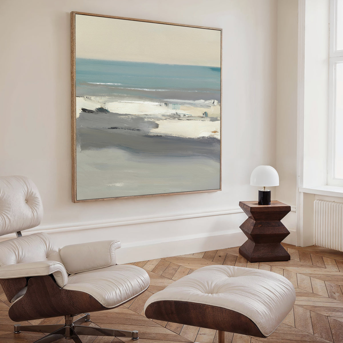 A modern, minimalist living room featuring a white leather lounge chair and ottoman, a wooden side table with a white lamp, and the Sandy Retreat Canvas Art - Square on the wall. Tall windows let in natural light for relaxing seaside vibes.