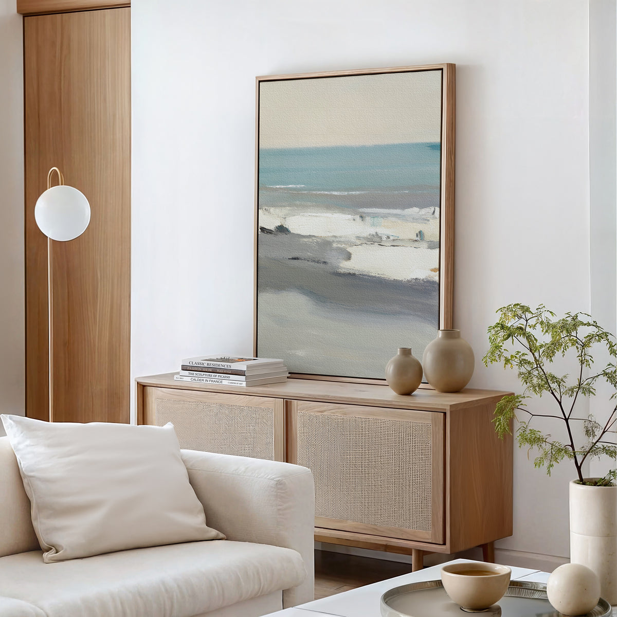 A minimalist living room featuring the Sandy Retreat Canvas Art - Vertical, a light wood sideboard, neutral vases, stacked books, a small plant, a white sofa with a cushion, and a modern globe floor lamp brings subtle seaside vibes.