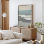 A minimalist living room featuring the Sandy Retreat Canvas Art - Vertical, a light wood sideboard, neutral vases, stacked books, a small plant, a white sofa with a cushion, and a modern globe floor lamp brings subtle seaside vibes.