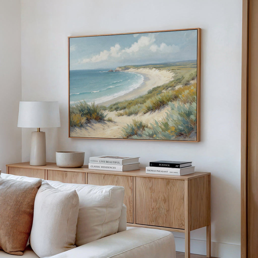 A cozy living room features a light wood sideboard, white table lamp, stacked books, and a decorative bowl. Above the light sofa hangs the Sandy Shores Canvas Art - Horizontal, bringing a serene coastal touch to the space.