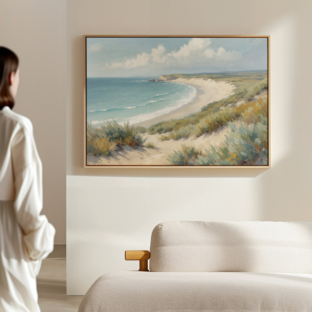 A woman stands in a softly lit room, admiring the Sandy Shores Canvas Art - Horizontal, which features a sandy beach and blue ocean with grassy dunes, hung above a beige upholstered bed on a light-colored wall.