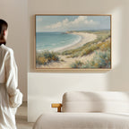 A woman stands in a softly lit room, admiring the Sandy Shores Canvas Art - Horizontal, which features a sandy beach and blue ocean with grassy dunes, hung above a beige upholstered bed on a light-colored wall.