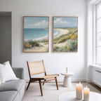 A bright living room features a modern chair, small table, lit candles, and Sandy Shores - Set of 2 large coastal canvases above the seating area, adding serene landscape decor to the crisp white wall.