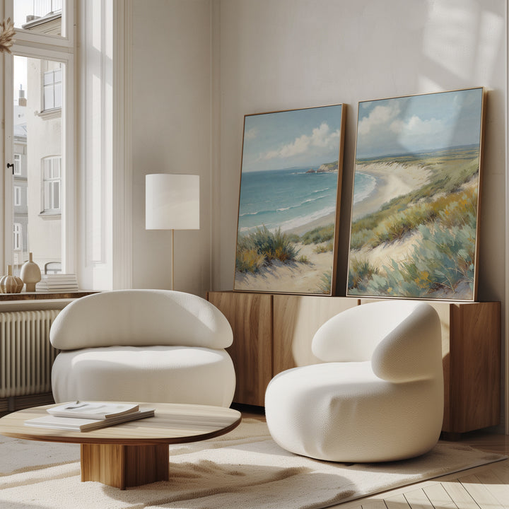 A modern living room features two white rounded chairs, a low wooden coffee table, and the "Sandy Shores - Set of 2" coastal canvas prints on a sideboard. Large windows fill the space with natural light for a bright, airy feel.
