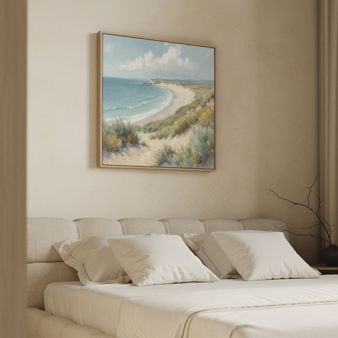 A cozy, neutral-toned bedroom showcases a large bed with white bedding and pillows, and the Sandy Shores Canvas Art - Square featuring sand dunes, grass, and ocean displayed above the headboard.