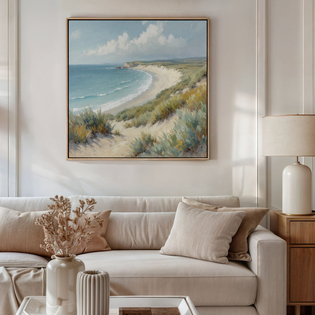 A cozy living room features a light beige sofa, neutral cushions, a lamp on a wooden side table, and a vase with dried flowers. Above the sofa hangs the Sandy Shores Canvas Art - Square, displaying a serene coastal landscape.