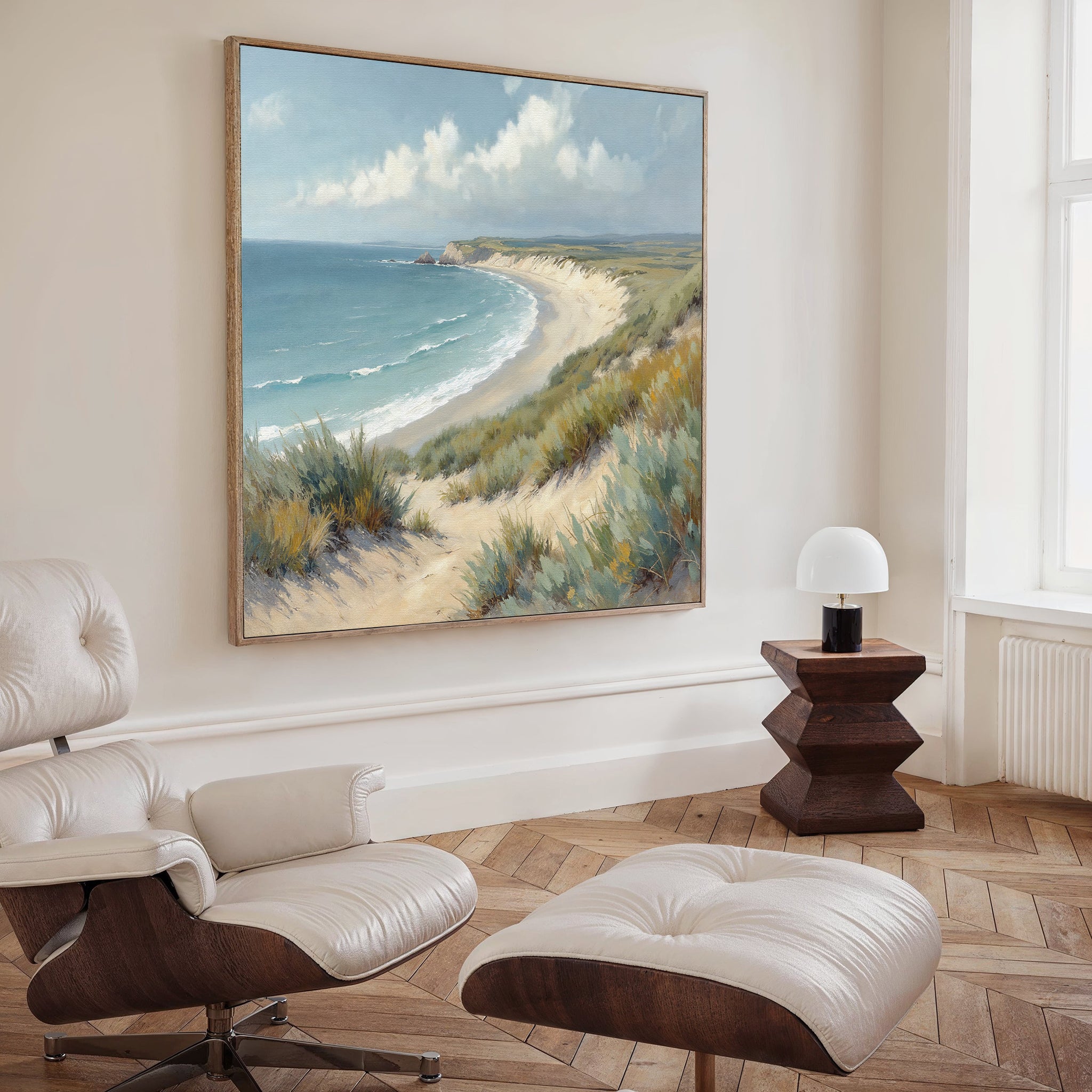 A modern living room with light wood floors, a white lounge chair and ottoman, a dark wood side table with a white lamp, and the Sandy Shores Canvas Art - Square featuring a striking coastal landscape on the wall.