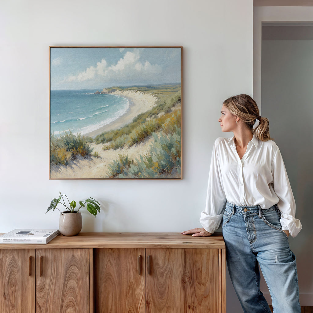 A woman in a white shirt and jeans stands by a wooden sideboard, admiring the Sandy Shores Canvas Art - Square on the wall. A plant and books rest nearby, enhancing the peaceful beach atmosphere of this display.