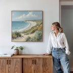 A woman in a white shirt and jeans stands by a wooden sideboard, admiring the Sandy Shores Canvas Art - Square on the wall. A plant and books rest nearby, enhancing the peaceful beach atmosphere of this display.