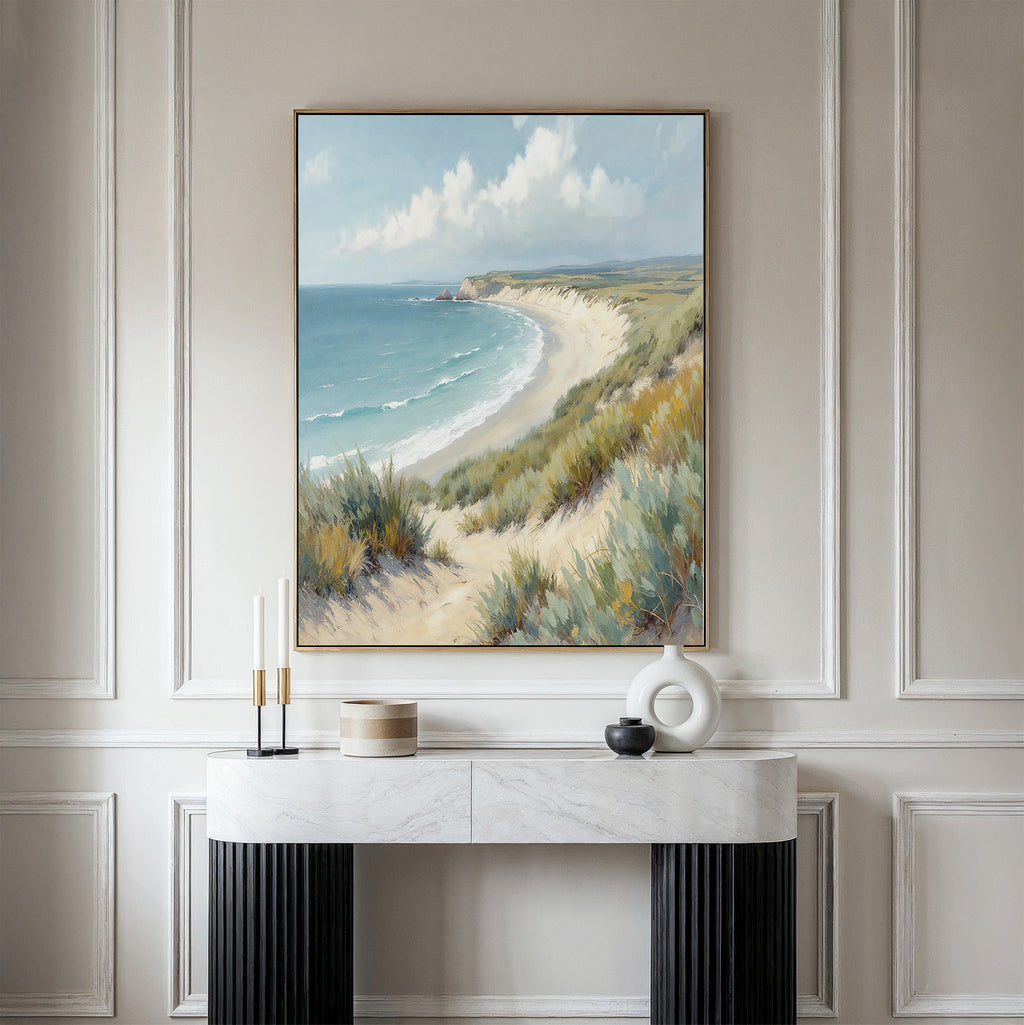 The Sandy Shores Canvas Art - Vertical featuring coastal dunes and a blue ocean hangs on a light beige wall above a marble console table adorned with candles and modern ceramics.