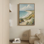 The Sandy Shores Canvas Art - Vertical, featuring a coastal scene with sandy beach, waves, and grassy dunes, hangs on a beige wall above a light sofa in a modern minimal living room. A shelf nearby holds books and a small decorative item.