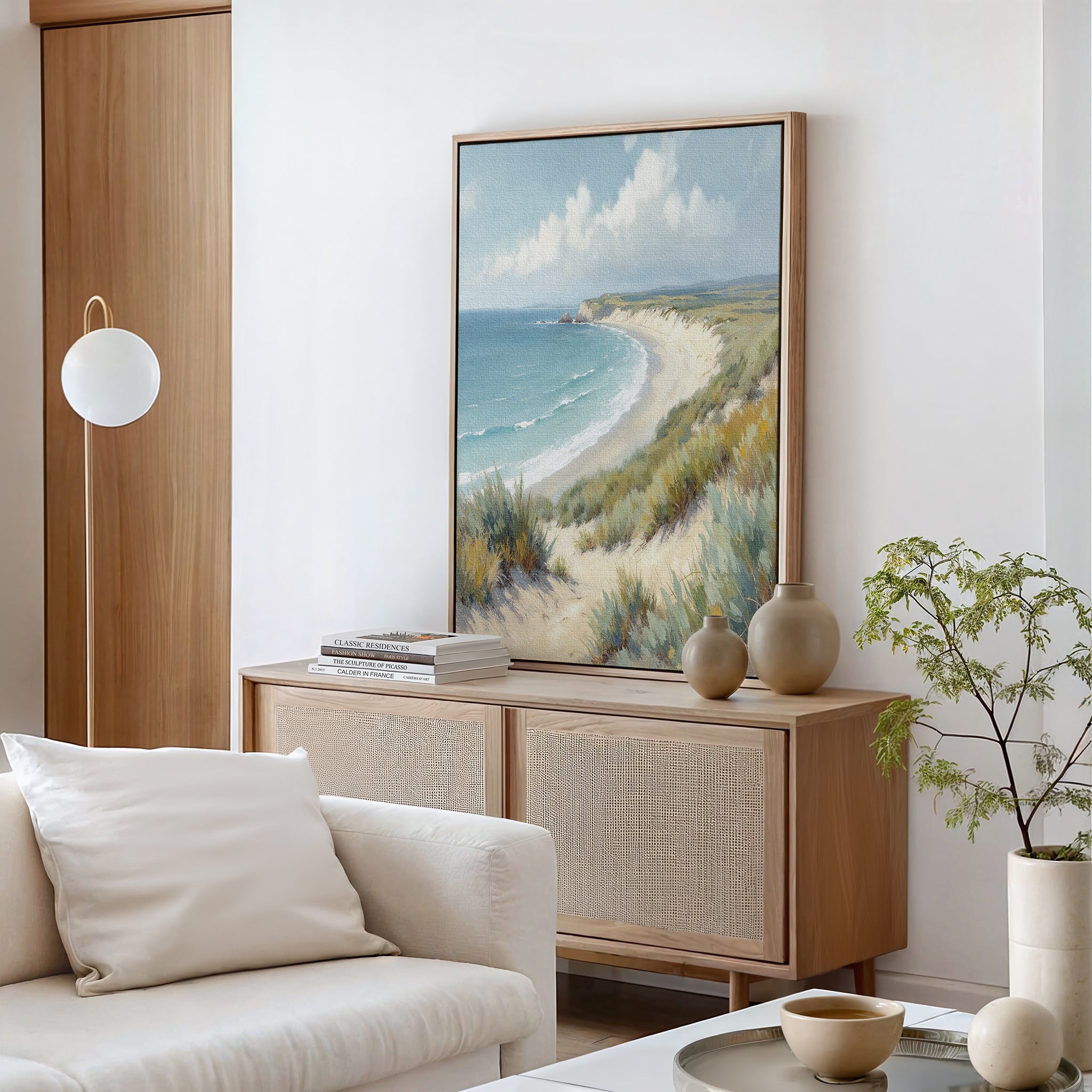 A coastal-themed living room features a light wood cabinet, beige vases, and the Sandy Shores Canvas Art - Vertical. A white sofa with a cushion and a modern globe lamp enhance the calm, airy vibe inspired by serene seaside escapes.