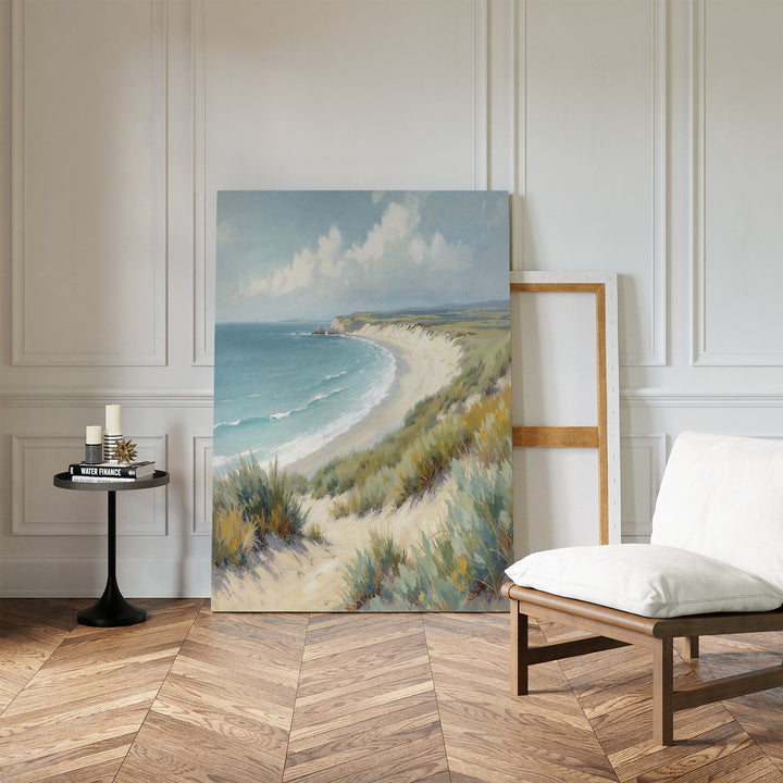 The Sandy Shores Canvas Art - Vertical rests against a white-paneled wall in a minimal room with a wooden chair, small round table with candles and books, and wood herringbone floor.