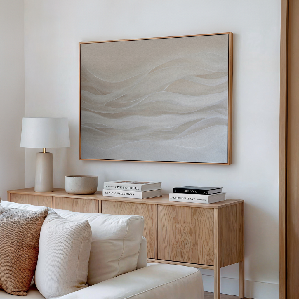 A modern living room features a light wooden sideboard with stacked books, a lamp, and neutral decor. Above, the Sandy Hues Abstract Canvas Art with soft beige and white waves brings sophisticated style to the space.