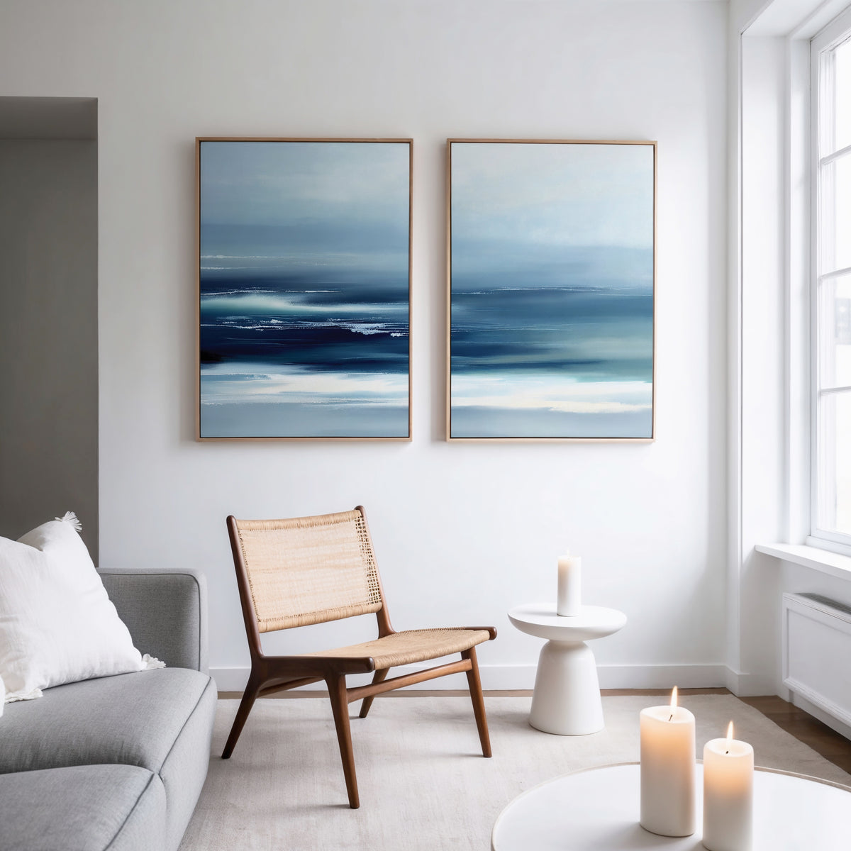A modern, minimalist living room with a light gray sofa, woven wooden chair, and tranquil wall art—Sapphire Current - Set Of 2 abstract prints above the chair. A small white side table holds a lit candle, and large windows bathe the space in natural light.