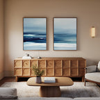 A modern living room features a wooden sideboard, a beige sofa, books on a wooden coffee table, and a small plant. The Sapphire Current - Set Of 2 wall art adds tranquility, while warm lighting creates a cozy atmosphere.