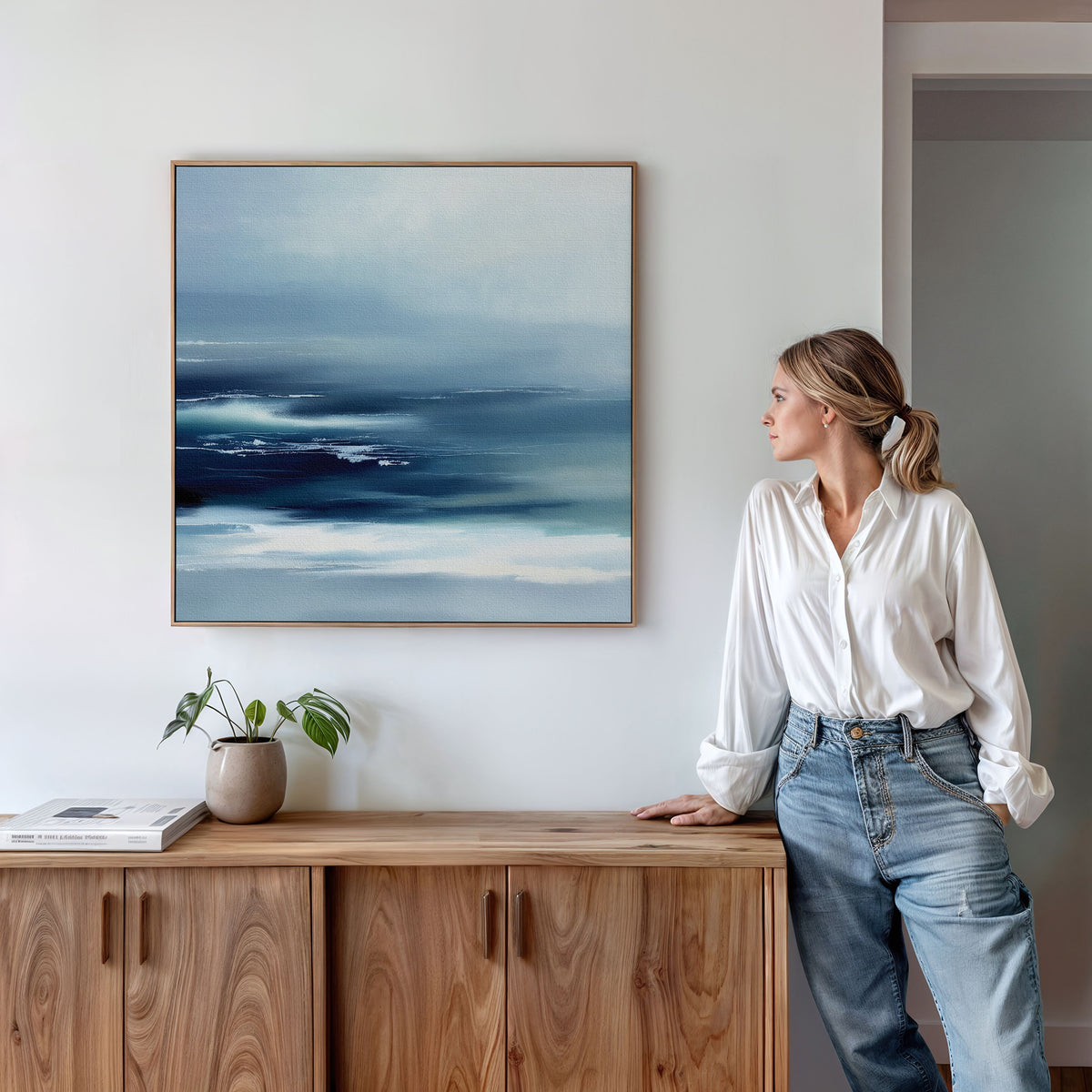 A woman in a white blouse and jeans stands by a wooden sideboard, admiring the Sapphire Current Canvas Art - Square. A potted plant and books complete the serene scene, emphasizing the calming effect of the tranquil wall art.