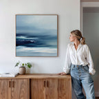 A woman in a white blouse and jeans stands by a wooden sideboard, admiring the Sapphire Current Canvas Art - Square. A potted plant and books complete the serene scene, emphasizing the calming effect of the tranquil wall art.
