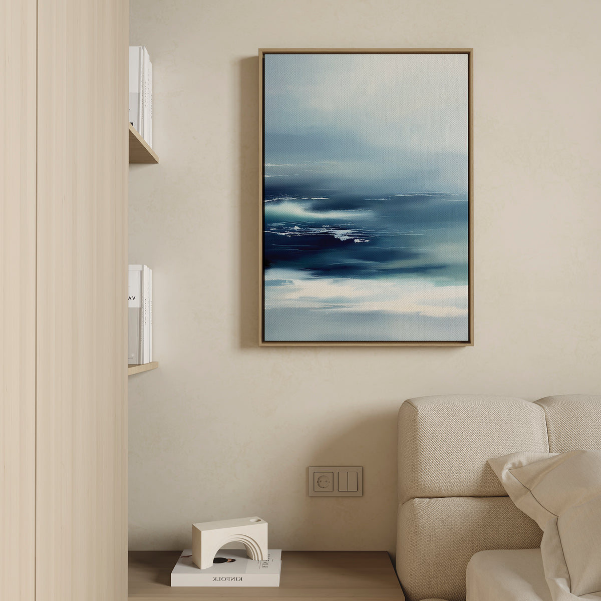 A modern living room with a light beige sofa, a wooden bookshelf topped with a white geometric decor, and tranquil wall art—a framed Sapphire Current Canvas Art - Vertical—hung on a cream-colored wall.