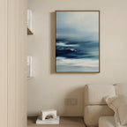 A modern living room with a light beige sofa, a wooden bookshelf topped with a white geometric decor, and tranquil wall art—a framed Sapphire Current Canvas Art - Vertical—hung on a cream-colored wall.