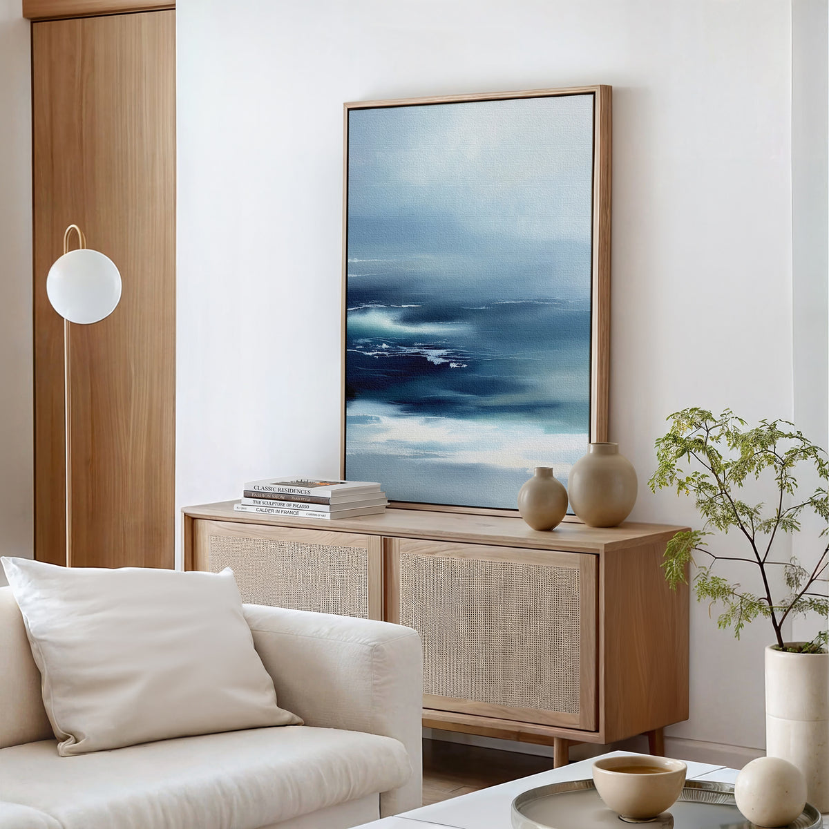 A modern living room features the Sapphire Current Canvas Art - Vertical, an abstract ocean painting in blue tones, above a light wood sideboard with vases and books. A white sofa, round light fixture, and potted plant complete the decor.