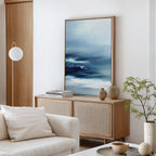 A modern living room features the Sapphire Current Canvas Art - Vertical, an abstract ocean painting in blue tones, above a light wood sideboard with vases and books. A white sofa, round light fixture, and potted plant complete the decor.