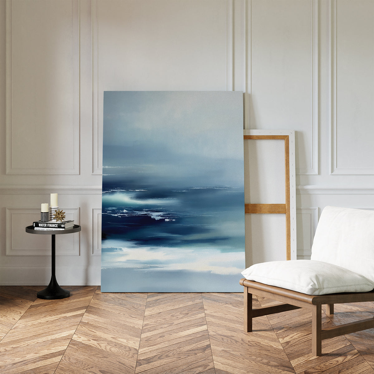 A modern room with herringbone wood floors features tranquil wall art—a Sapphire Current Canvas Art - Vertical print of the ocean—leaning against a white paneled wall, beside an empty frame, a cushioned chair, and a small round table with candles and books.
